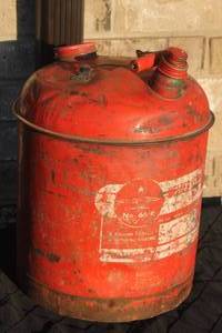 lot 27 image: Vintage red 5-gallon metal gas can featuring weathered graphics and a wooden handle grip.