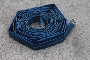 lot 34 image: Goodyear Spiraflex blue flat discharge hose coiled on concrete, showing attached hose couplings and light surface wear.