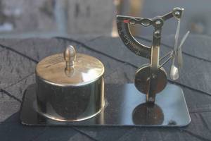 lot 40 image: Vintage brass postal scale desktop set mounted on a black base, featuring a polished cylindrical stamp holder or inkwell and a miniature balance scale.